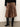 A person wearing the Sanctuary Clothing nostalgia pleated midi skirt in mocha leo, paired with knee-high black leather boots and a black top, stands on a gray floor against an off-white wall.