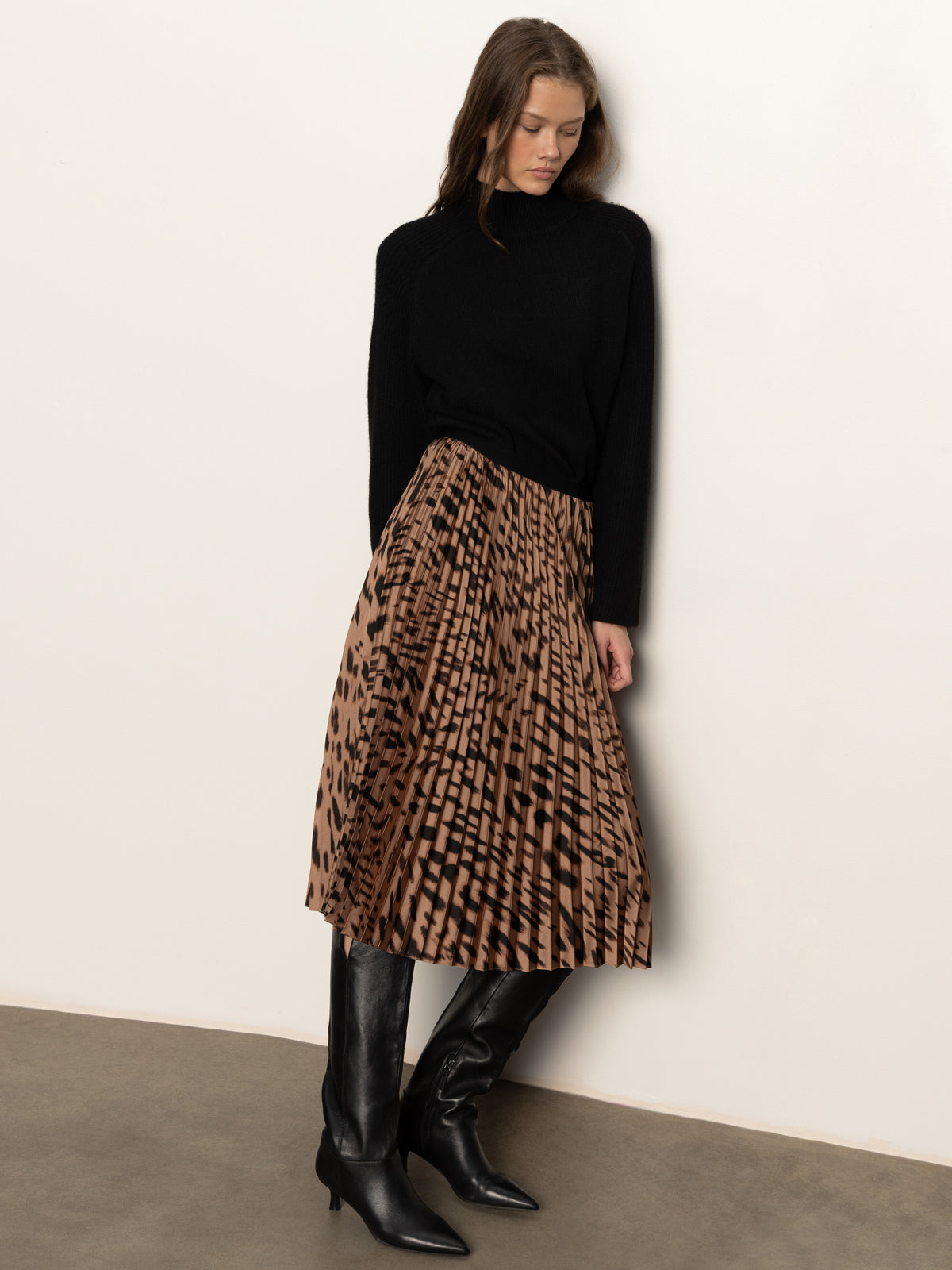 A woman stands against a plain wall wearing Sanctuary Clothing's nostalgia pleated midi skirt in mocha leo, paired with a black sweater and knee-high black boots. She looks down with her hands by her sides.