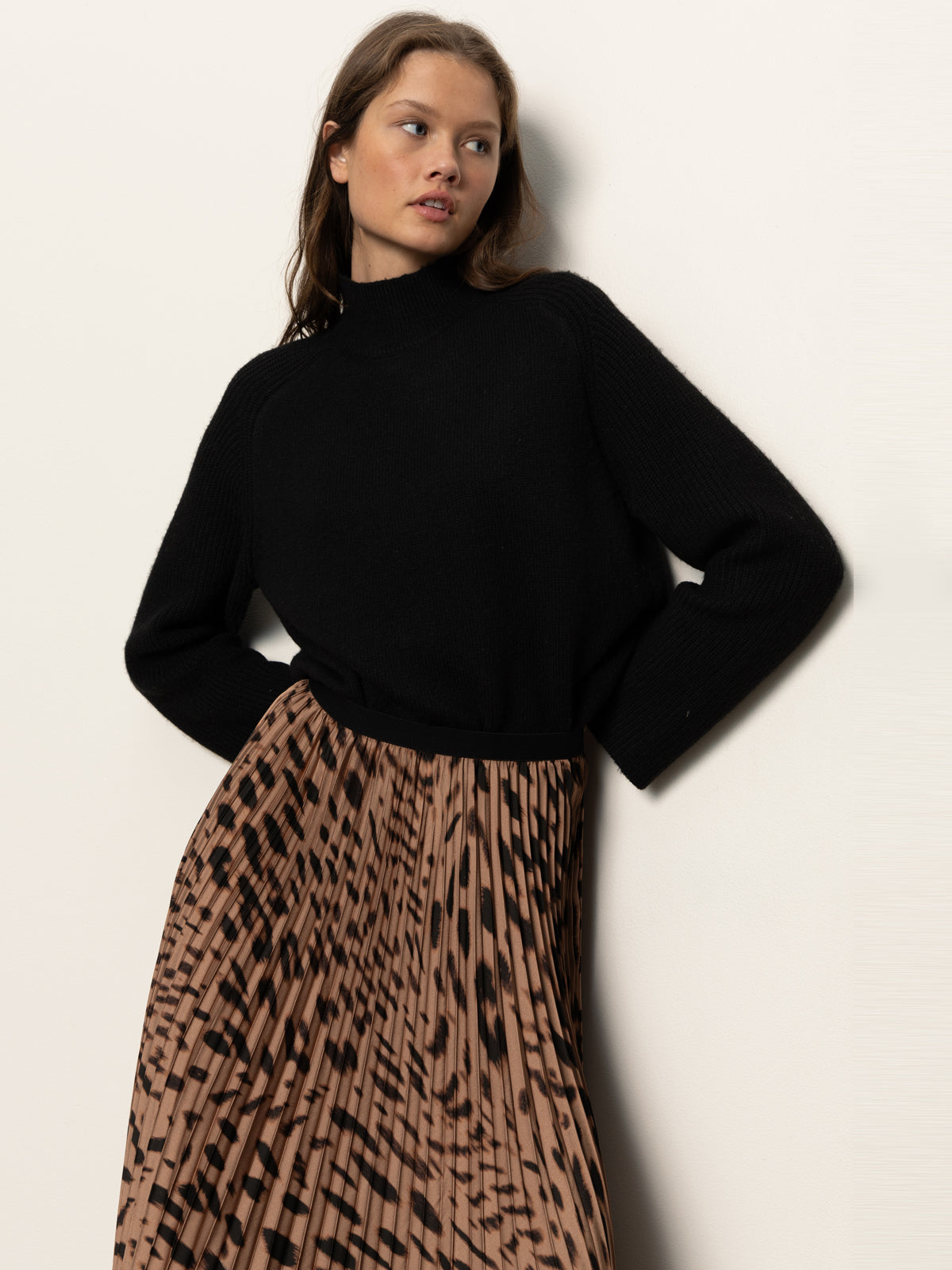 A woman stands against a light background, wearing a black long-sleeve sweater and the Sanctuary Clothing nostalgia pleated midi skirt in mocha leo, featuring a brown and black abstract pattern, looking to the side with one hand on her hip.