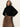 A woman stands against a light background, wearing a black long-sleeve sweater and the Sanctuary Clothing nostalgia pleated midi skirt in mocha leo, featuring a brown and black abstract pattern, looking to the side with one hand on her hip.