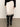 A person wears the Sanctuary Clothing evermore pleated midi skirt in light oat with black lace trim, styled with a black sweater and black knee-high boots, standing against a light background.