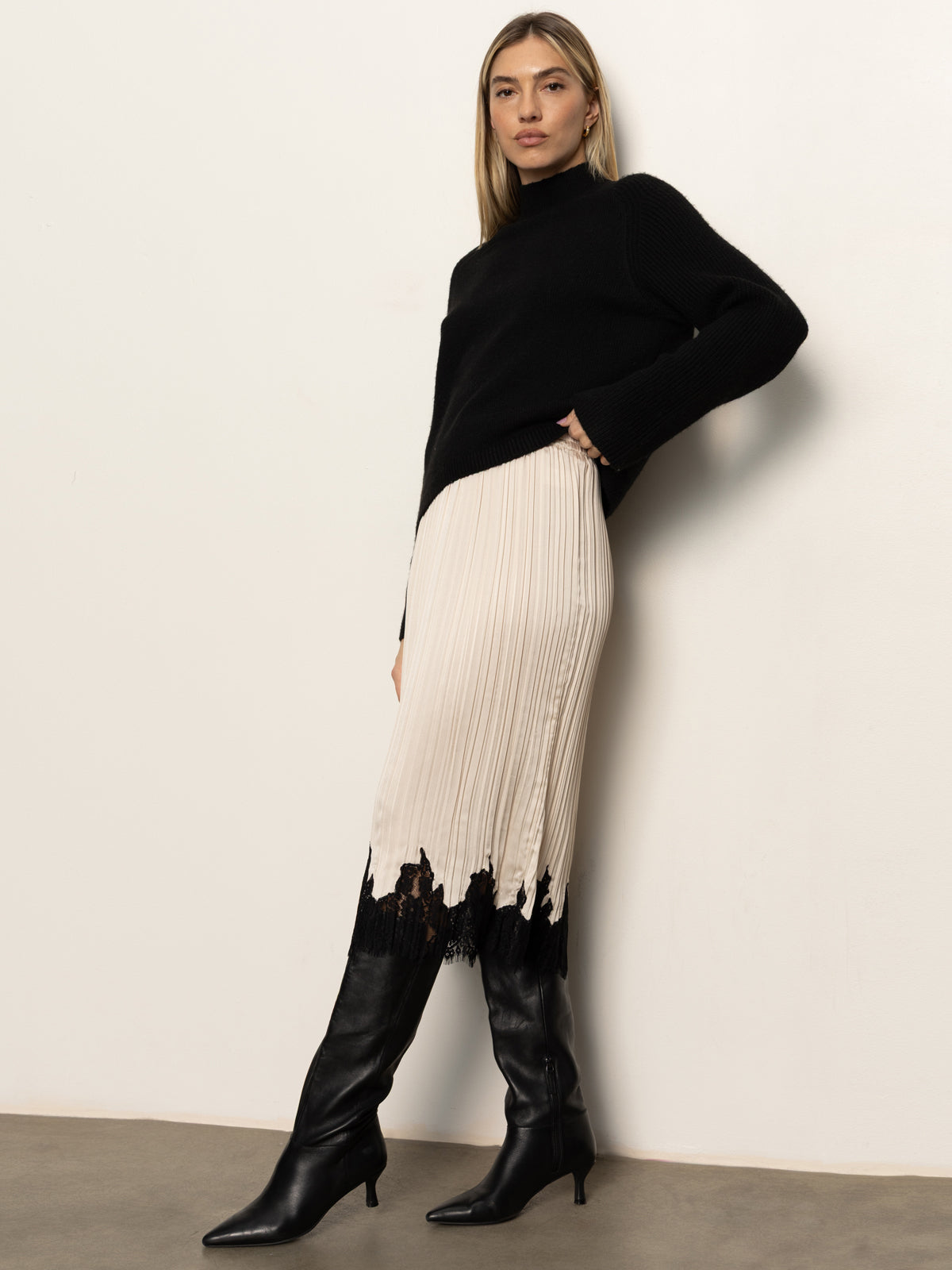 A woman with straight light brown hair and a neutral expression stands against a white wall wearing Sanctuary Clothing's evermore pleated midi skirt in light oat, styled with a black turtleneck sweater and black knee-high heeled boots.