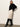 A woman with straight light brown hair and a neutral expression stands against a white wall wearing Sanctuary Clothing's evermore pleated midi skirt in light oat, styled with a black turtleneck sweater and black knee-high heeled boots.