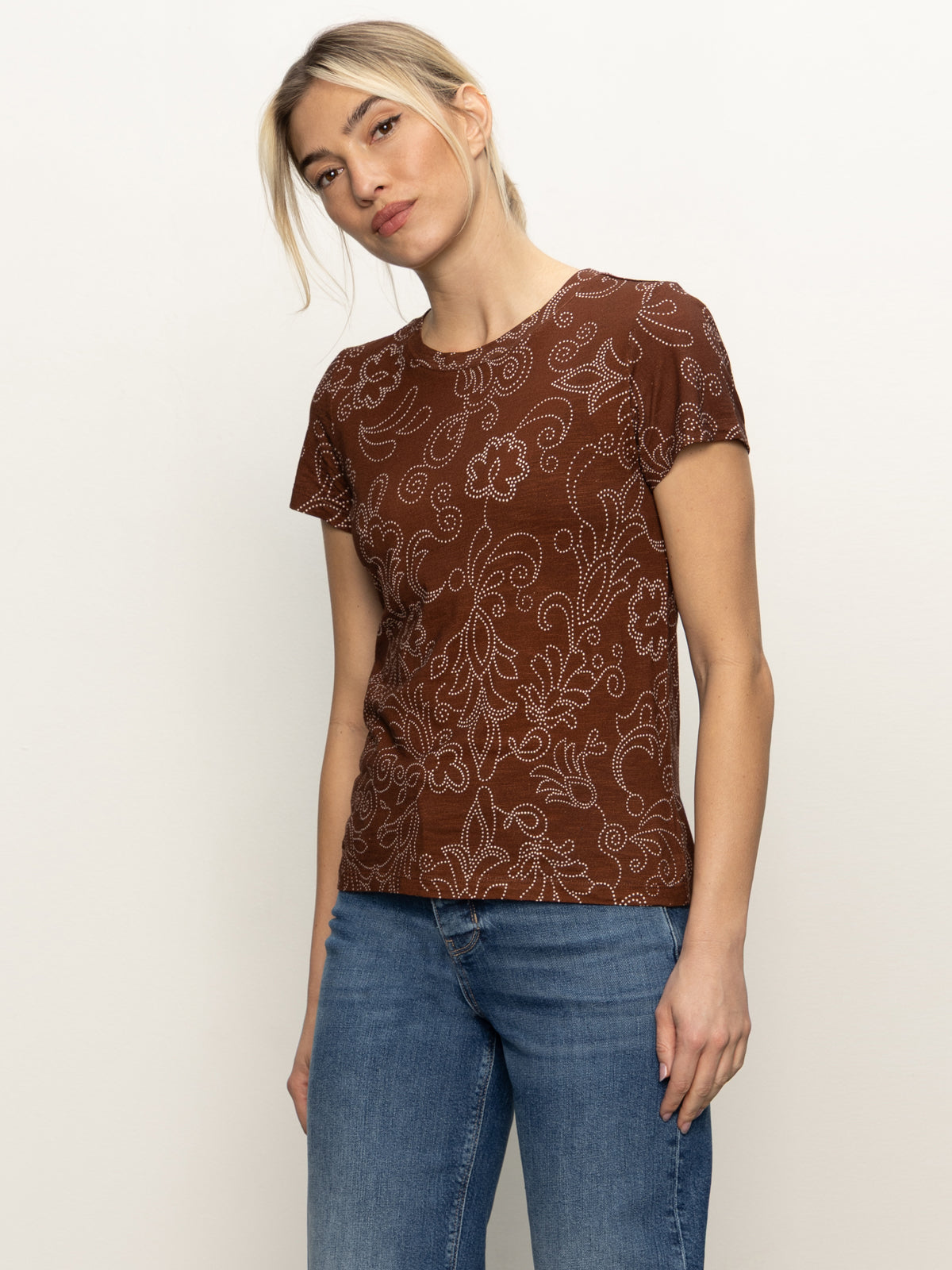 A woman models the Sanctuary Clothing "the perfect tee boheme," a brown short-sleeve shirt with a white floral pattern, paired with blue jeans, standing against a plain light background and gazing to the side.