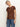 A woman models the Sanctuary Clothing "the perfect tee boheme," a brown short-sleeve shirt with a white floral pattern, paired with blue jeans, standing against a plain light background and gazing to the side.