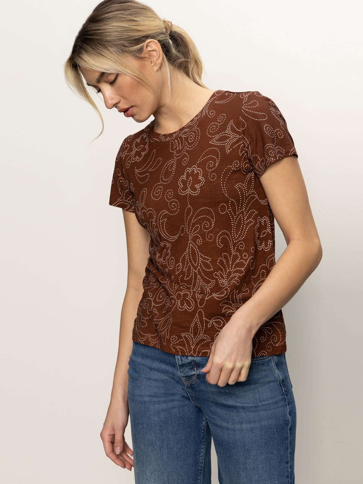 A woman with blonde hair in a ponytail wears Sanctuary Clothing’s "the perfect tee boheme," a brown short-sleeve shirt with a white floral pattern, paired with blue jeans. She stands against a plain light background, looking down.