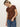 A woman with blonde hair in a ponytail wears Sanctuary Clothing’s "the perfect tee boheme," a brown short-sleeve shirt with a white floral pattern, paired with blue jeans. She stands against a plain light background, looking down.
