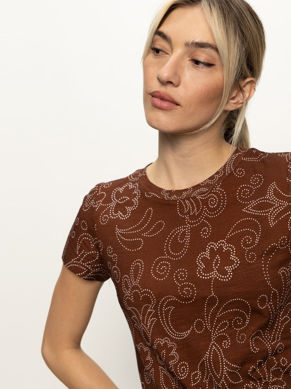 A woman with blonde hair in a low ponytail wears the Sanctuary Clothing "the perfect tee boheme," a brown short-sleeve shirt with a white dotted floral and swirl pattern, looking slightly left against a plain light background.