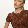 A woman with blonde hair in a low ponytail wears the Sanctuary Clothing "the perfect tee boheme," a brown short-sleeve shirt with a white dotted floral and swirl pattern, looking slightly left against a plain light background.