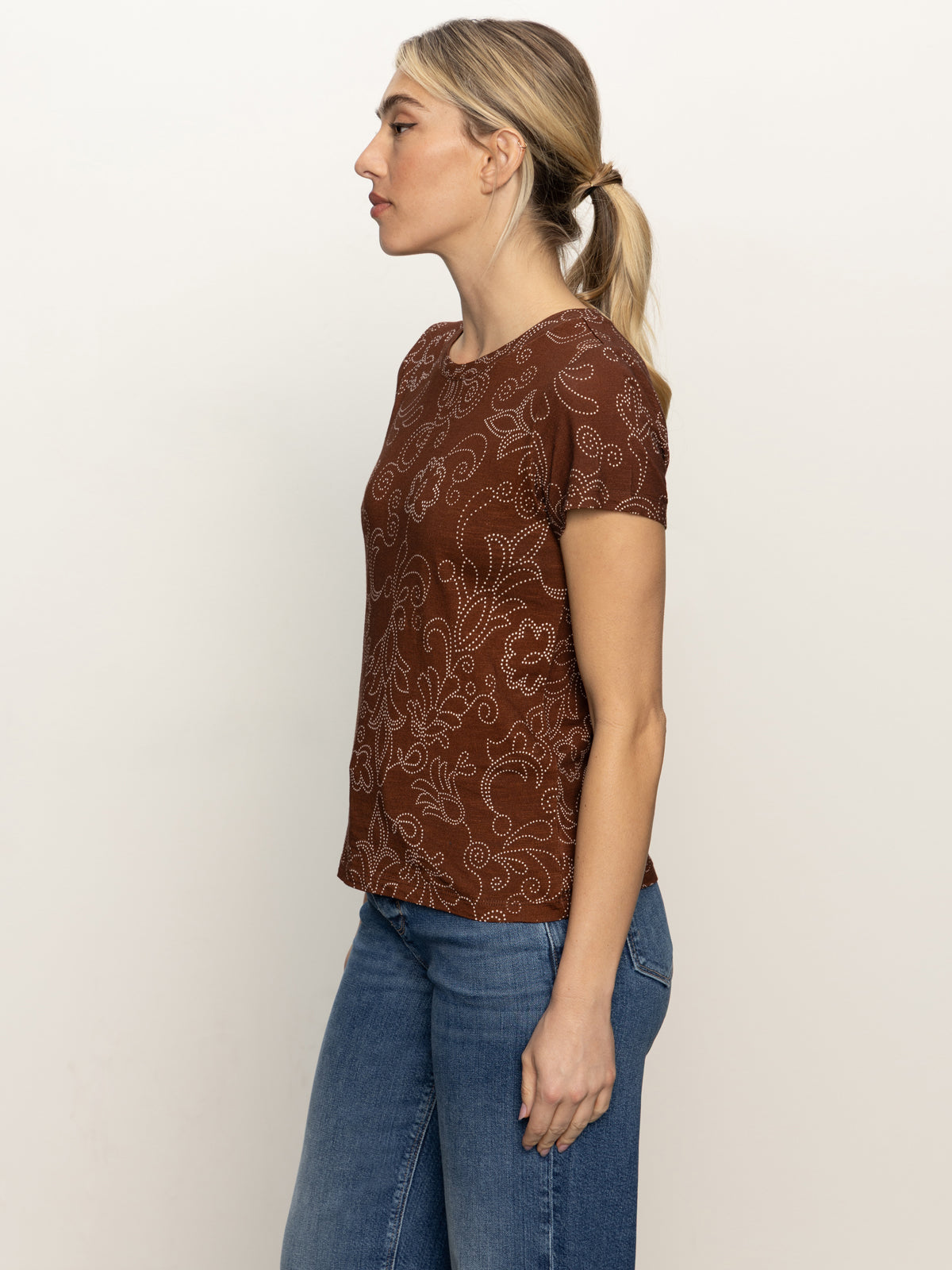 A woman with blonde hair in a low ponytail stands in profile against a plain background, wearing Sanctuary Clothing's the perfect tee boheme and blue jeans.