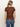 A woman with blonde hair in a low ponytail is seen from behind, wearing the perfect tee boheme by Sanctuary Clothing—a brown short-sleeve top with a subtle floral pattern—paired with blue jeans against a plain light background.