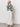 A woman stands against a white wall wearing Sanctuary Clothing's the perfect tee in green oasis, paired with high-waisted white wide-leg pants and black sandals. She places her hands in her pockets, gazing slightly upward.
