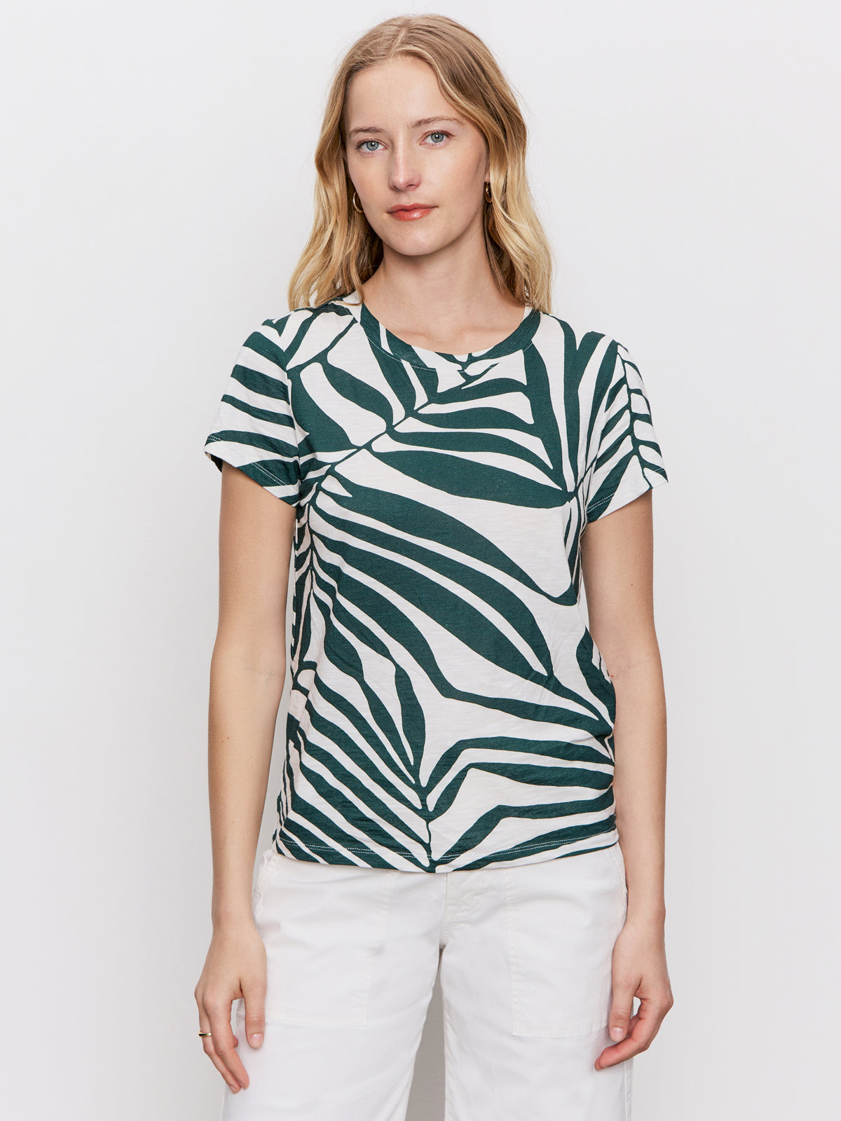 A woman with blonde hair wears Sanctuary Clothing's "the perfect tee green oasis"—a green and white leaf-patterned short-sleeve shirt—paired with white pants, standing against a plain white background and looking neutrally at the camera.