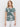 A woman with blonde hair wears Sanctuary Clothing's "the perfect tee green oasis"—a green and white leaf-patterned short-sleeve shirt—paired with white pants, standing against a plain white background and looking neutrally at the camera.