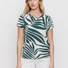 A woman with blonde hair wears Sanctuary Clothing's "the perfect tee green oasis"—a green and white leaf-patterned short-sleeve shirt—paired with white pants, standing against a plain white background and looking neutrally at the camera.