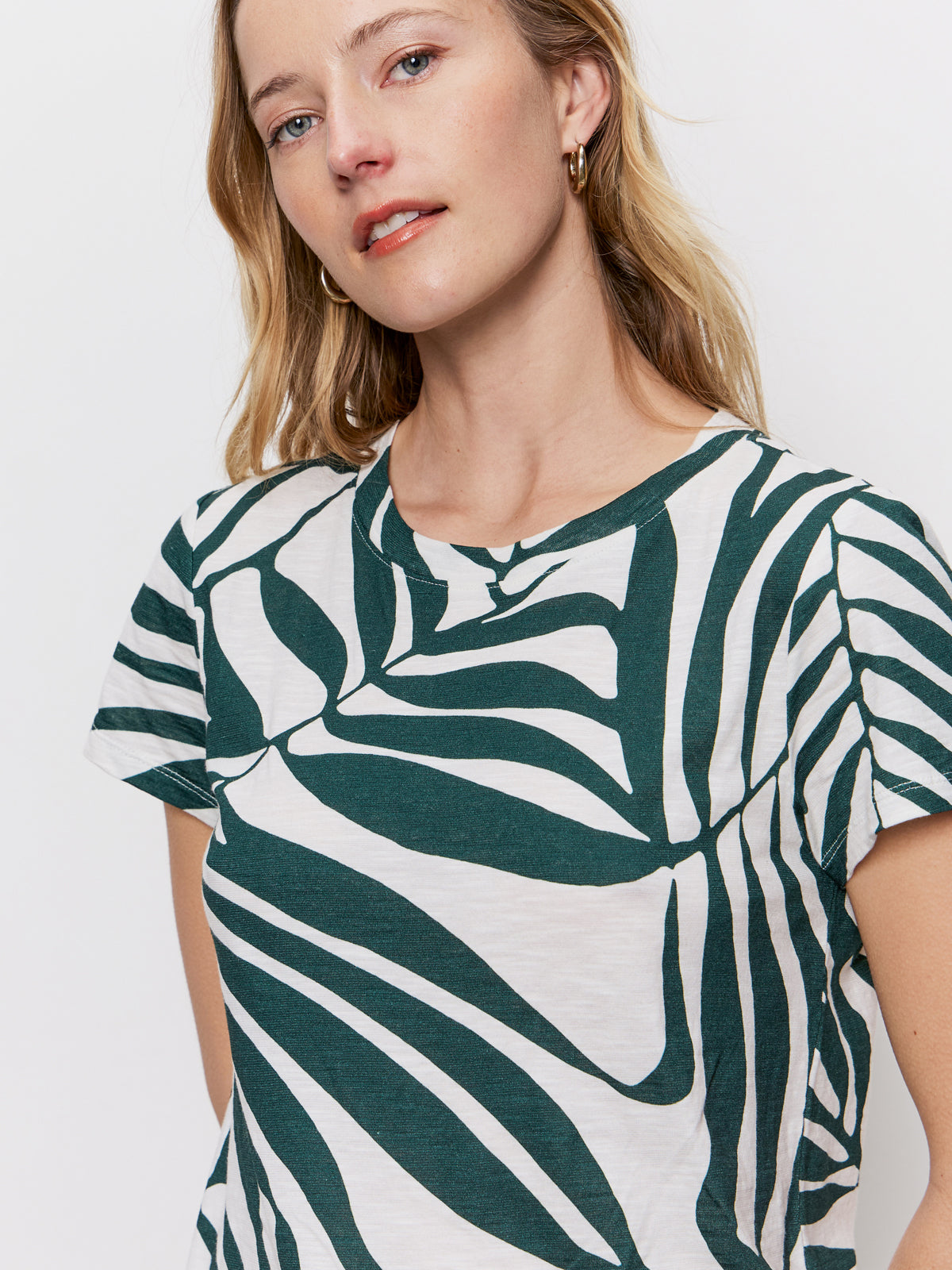 A woman with light skin and blonde hair wears the Sanctuary Clothing "the perfect tee green oasis"—a white T-shirt featuring a bold green abstract pattern. She stands against a plain white background, smiling gently to the side.