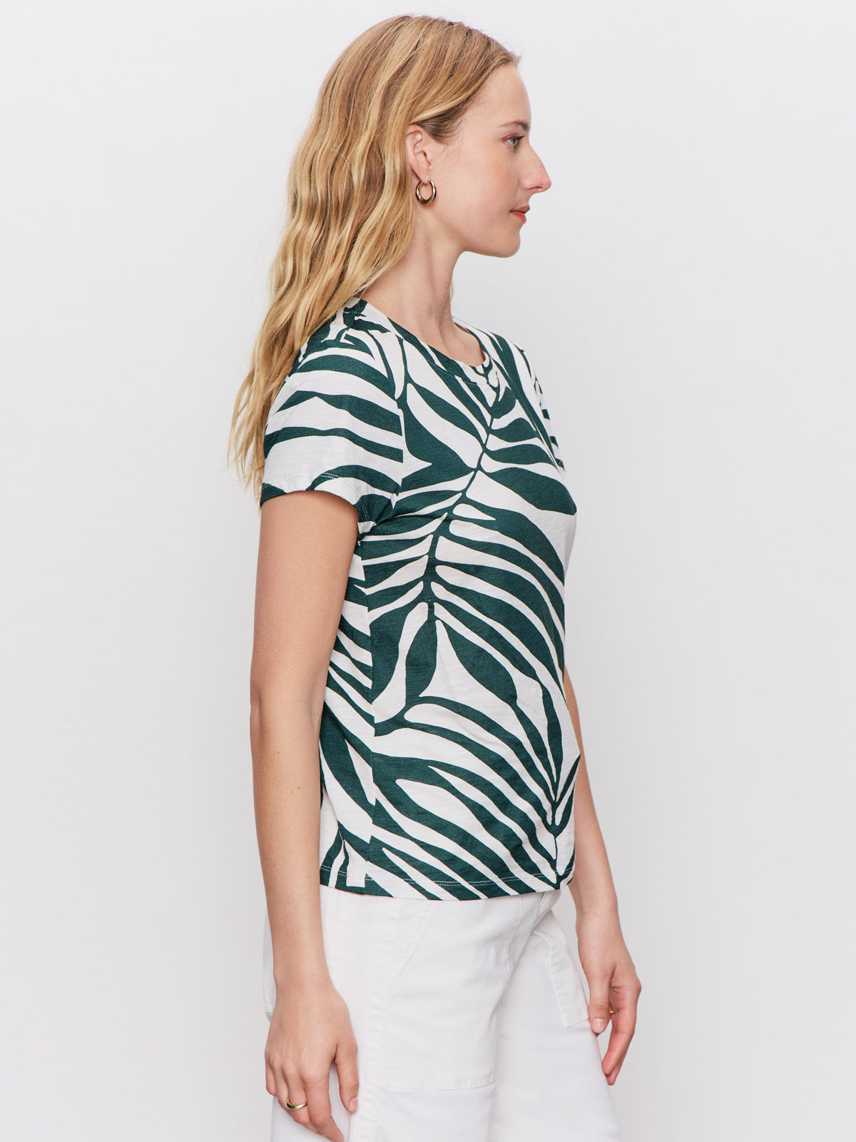 A woman with wavy blonde hair stands in profile against a plain white background, wearing Sanctuary Clothing's "the perfect tee green oasis"—a short-sleeve white t-shirt with a bold green leaf pattern—and white pants.