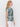A woman with wavy blonde hair stands in profile against a plain white background, wearing Sanctuary Clothing's "the perfect tee green oasis"—a short-sleeve white t-shirt with a bold green leaf pattern—and white pants.