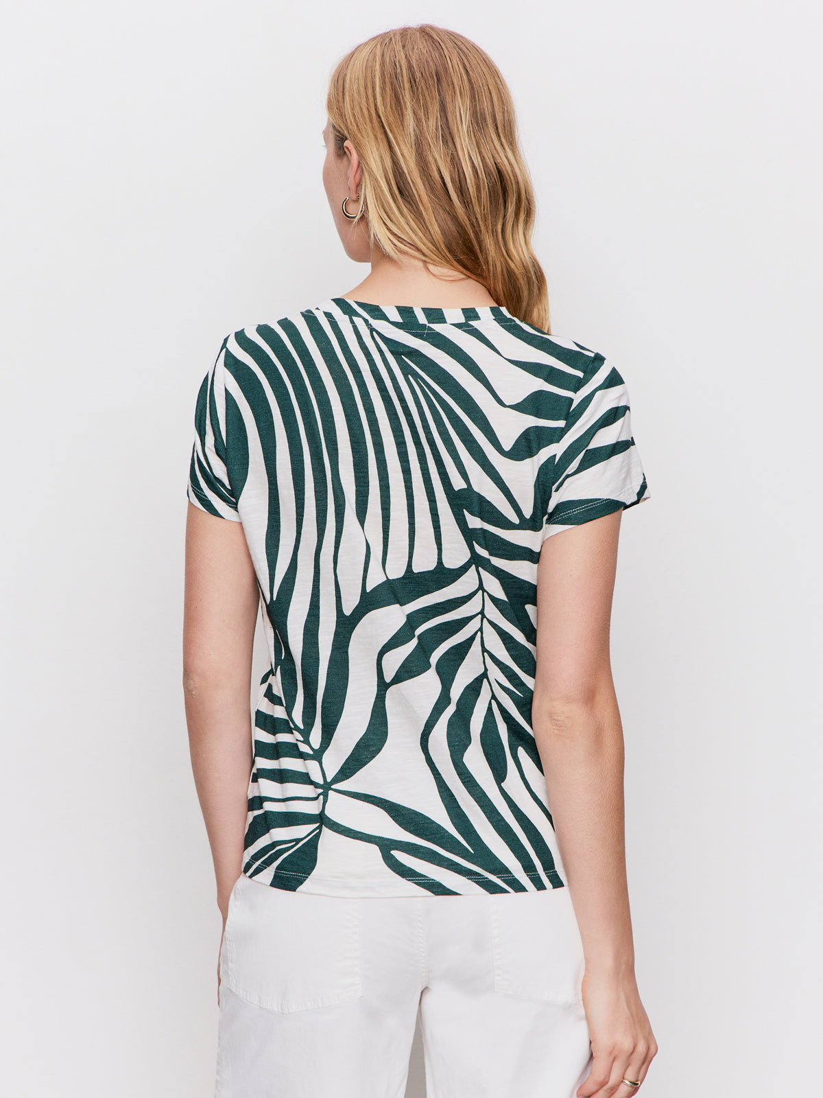 A woman with light brown hair is seen from the back wearing Sanctuary Clothing's "the perfect tee green oasis," featuring a bold dark green abstract leaf pattern, paired with white shorts against a plain white background.
