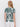 A woman with light brown hair is seen from the back wearing Sanctuary Clothing's "the perfect tee green oasis," featuring a bold dark green abstract leaf pattern, paired with white shorts against a plain white background.