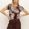 A woman with straight blonde hair stands against a plain background wearing Sanctuary Clothing's "the perfect tee mesa palm," a maroon t-shirt with beige tropical leaf prints, paired with matching maroon shorts.