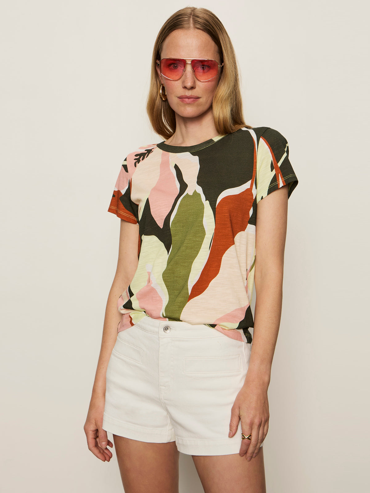 A woman stands confidently against a light background, wearing Sanctuary Clothing’s "the perfect tee reef vibes" with a colorful abstract pattern, paired with pink sunglasses and white shorts, her hand casually in her pocket.