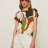 A woman stands confidently against a light background, wearing Sanctuary Clothing’s "the perfect tee reef vibes" with a colorful abstract pattern, paired with pink sunglasses and white shorts, her hand casually in her pocket.
