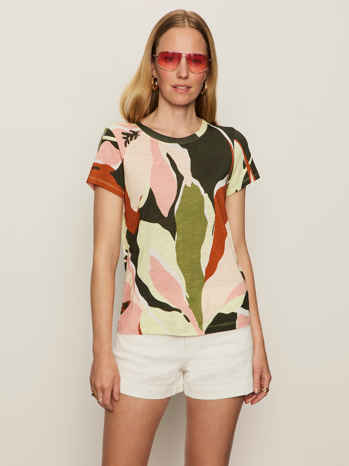 A woman in Sanctuary Clothing’s “the perfect tee reef vibes”—a colorful leaf-patterned short-sleeve shirt—paired with white shorts and pink sunglasses, stands against a plain light background.