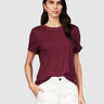 A woman with long dark hair models the Sanctuary Clothing perfect tee remix night berry, a maroon short-sleeve t-shirt, paired with white high-waisted pants against a plain light gray background, one hand in her pocket.
