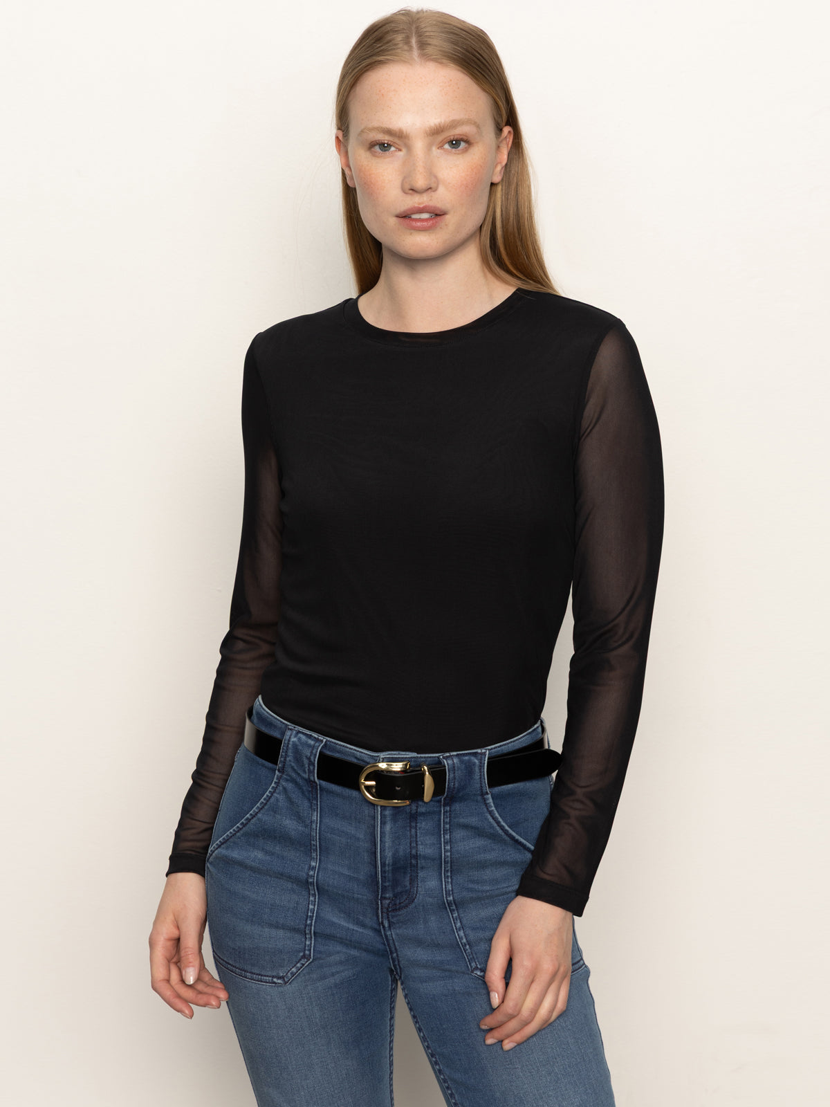 A woman with straight, light brown hair wears the Sanctuary Clothing long sleeveperfect mesh tee in black, styled with blue high-waisted jeans and a black belt with a gold buckle, standing against a plain light background.