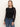 A woman with straight, light brown hair wears the Sanctuary Clothing long sleeveperfect mesh tee in black, styled with blue high-waisted jeans and a black belt with a gold buckle, standing against a plain light background.