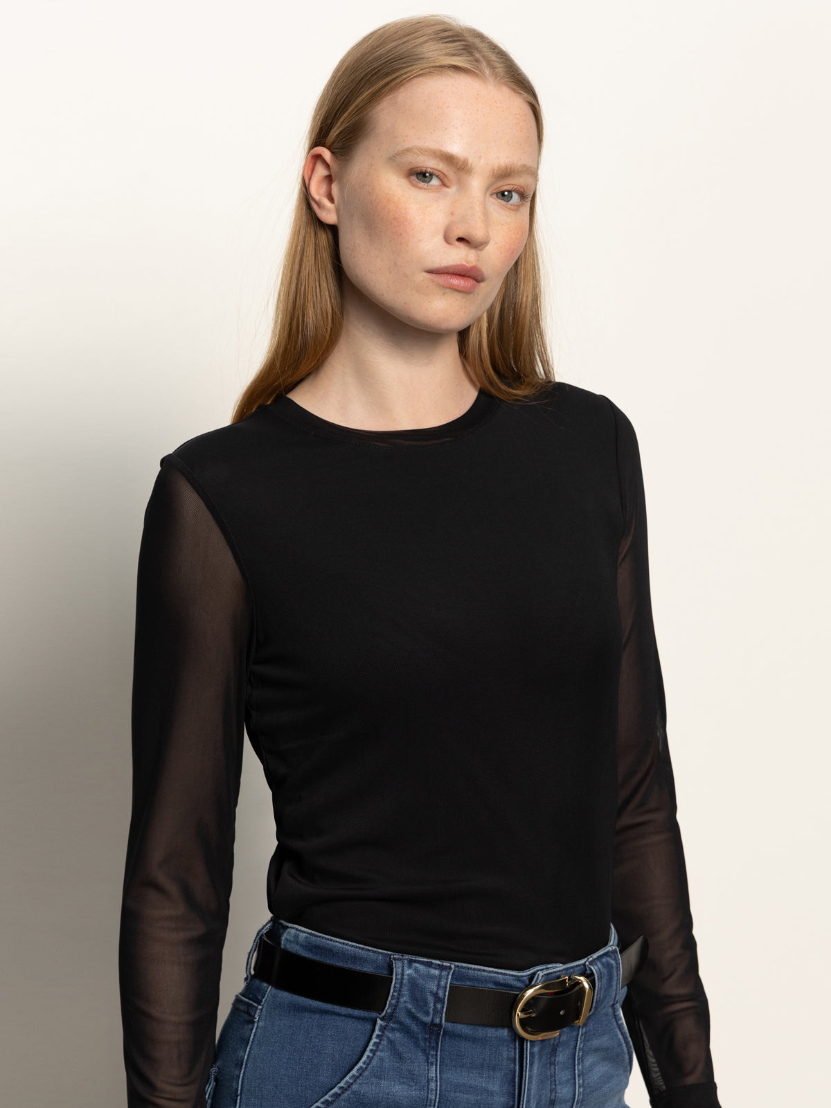A person with long, straight blond hair wears the Sanctuary Clothing long sleeveperfect mesh tee in black, styled with blue jeans and a black belt, standing against a neutral background.