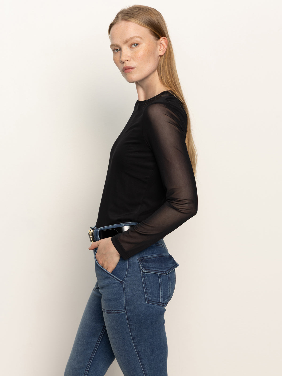 A woman with long, straight blonde hair stands in profile with hands in her pockets, wearing Sanctuary Clothing's black long sleeveperfect mesh tee and blue jeans against a plain light background.
