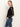 A woman with long, straight blonde hair stands in profile with hands in her pockets, wearing Sanctuary Clothing's black long sleeveperfect mesh tee and blue jeans against a plain light background.