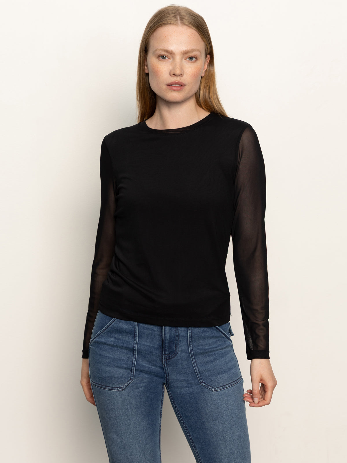 A woman with straight, light brown hair wears the Sanctuary Clothing long sleeveperfect mesh tee in black—featuring fitted sheer sleeves—paired with blue jeans, and stands against a plain light background.