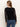 A woman with long, straight light brown hair stands with her back to the camera, wearing the Sanctuary Clothing long sleeveperfect mesh tee in black and blue jeans against a light plain background.