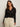 A woman with long brown hair wears the Sanctuary Clothing pucker up dreamgirl black textured button-up cardigan with a V-neck, paired with cream pants, standing against a plain off-white background and looking confidently at the camera.