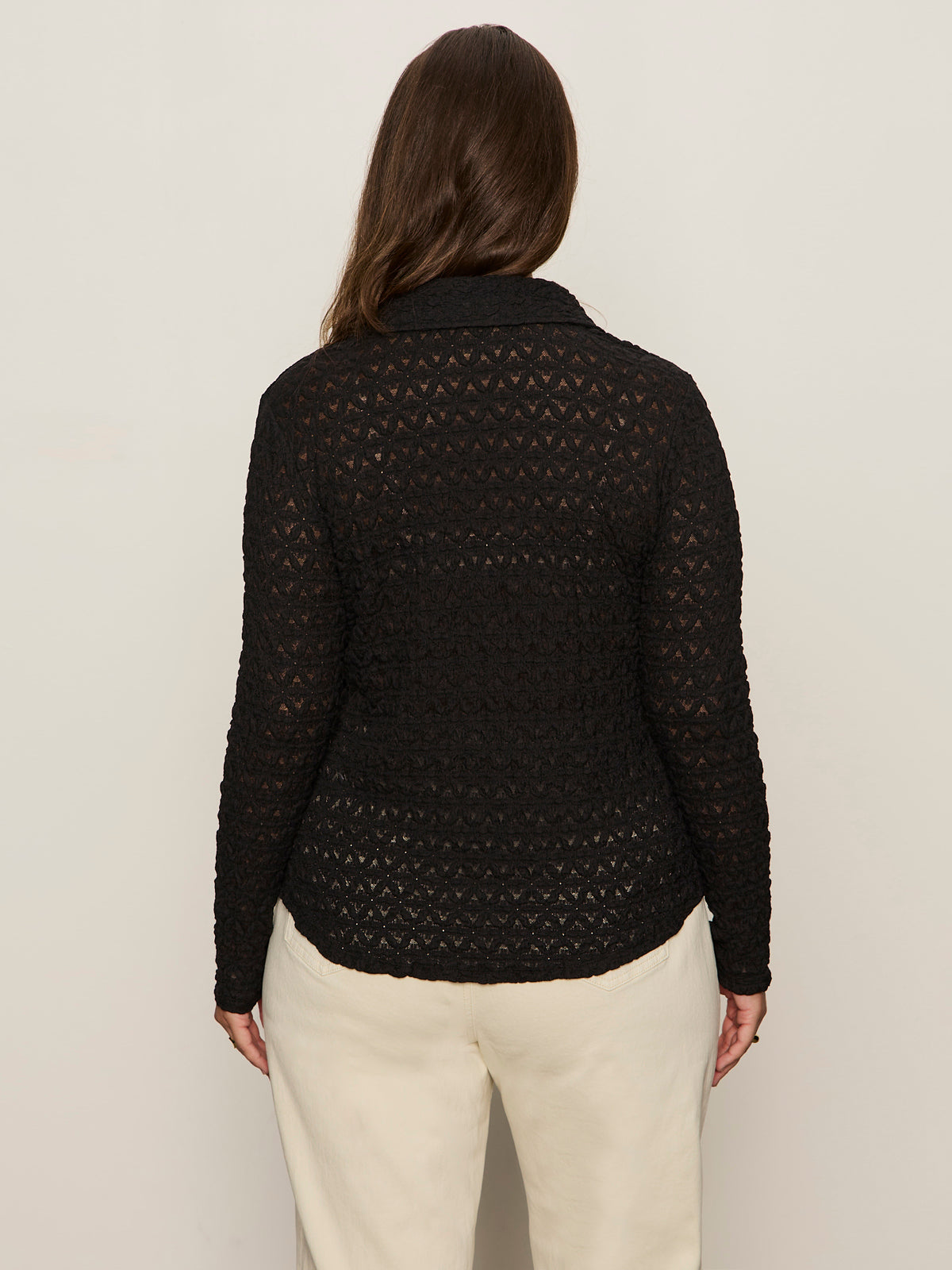 A person with long brown hair is seen from the back, wearing Sanctuary Clothing’s pucker up dreamgirl black textured long-sleeve top and cream pants, standing against a plain light background.