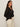 A woman with long brown hair models the Sanctuary Clothing "pucker up dreamgirl black" textured long-sleeve top and off-white pants, standing sideways and smiling slightly at the camera against a plain light background.