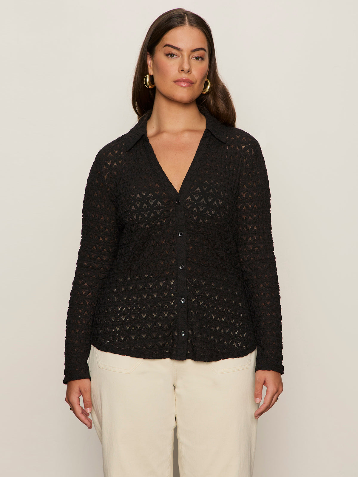 A woman with long brown hair wears the Sanctuary Clothing pucker up dreamgirl black long-sleeve lace shirt with a collar and cream-colored pants, standing against a plain light background.