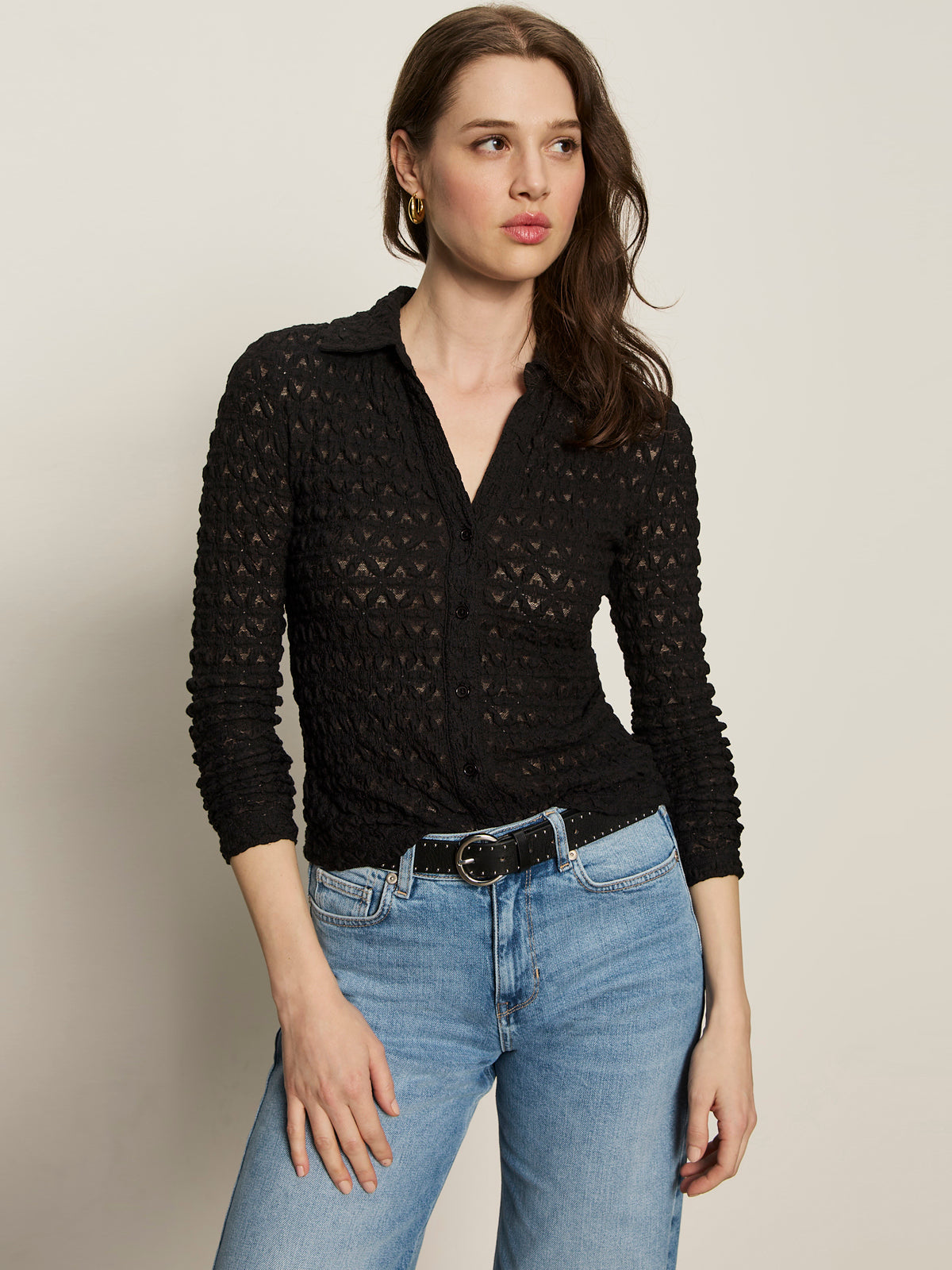 A woman with long brown hair wears the Sanctuary Clothing dreamgirl lace button up black blouse, styled with light blue jeans and a black belt. She stands against a plain light background, looking slightly to the side.