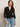 A woman with long brown hair wears the Sanctuary Clothing dreamgirl lace button up black blouse, styled with light blue jeans and a black belt. She stands against a plain light background, looking slightly to the side.
