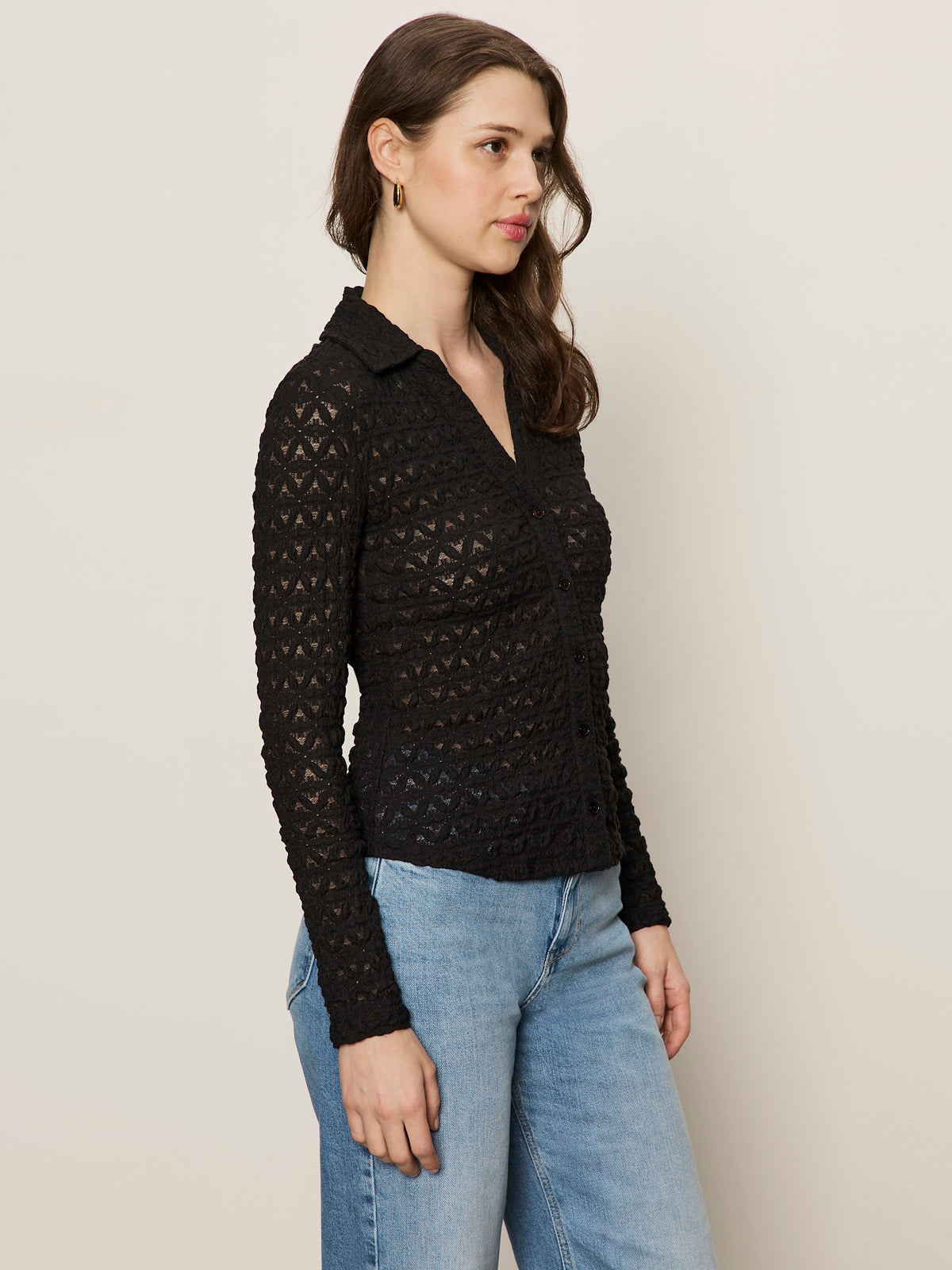 A woman stands in profile against a plain background, wearing the Sanctuary Clothing dreamgirl lace button up black shirt with a collar and light blue jeans. She has loose brown hair and small hoop earrings.