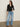 A woman with long brown hair and a neutral expression stands against a plain wall wearing the Sanctuary Clothing "dreamgirl lace button up" black top, paired with light blue high-waisted wide-leg jeans and black open-toe heeled sandals.