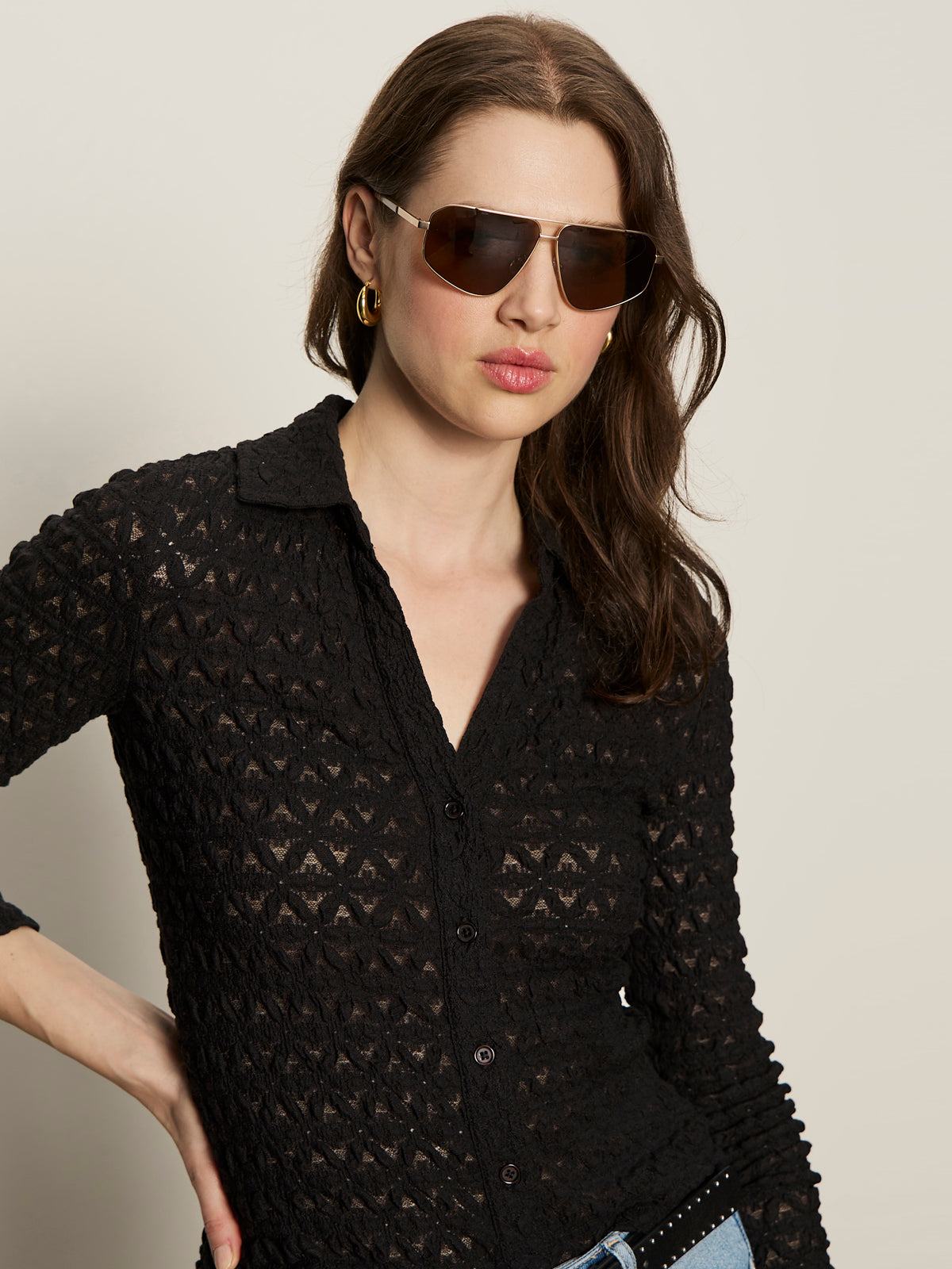 A woman with long brown hair, large sunglasses, and gold hoop earrings wears the Sanctuary Clothing dreamgirl lace button up black shirt, posing with one hand on her hip against a plain background.