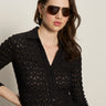 A woman with long brown hair, large sunglasses, and gold hoop earrings wears the Sanctuary Clothing dreamgirl lace button up black shirt, posing with one hand on her hip against a plain background.