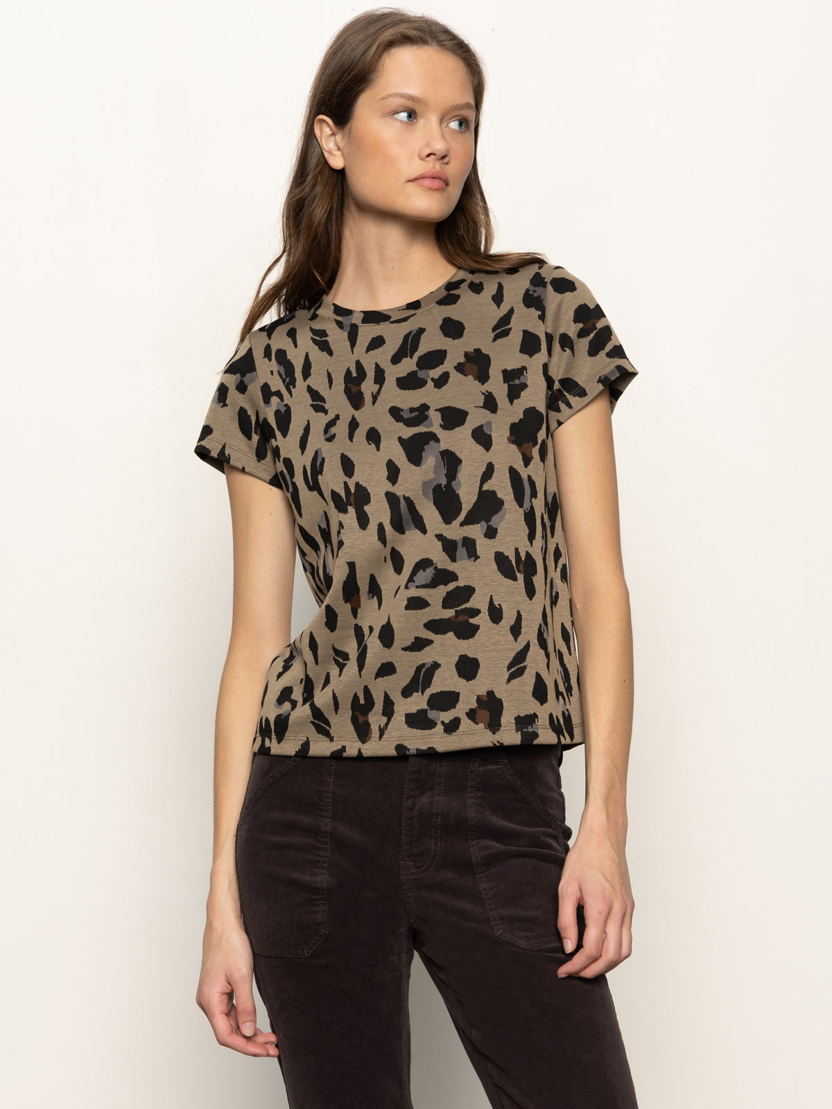 A woman with long brown hair wears the Sanctuary Clothing double knit perfect tee in winter leopard and dark pants, standing against an off-white background. She looks to the side with a neutral expression.