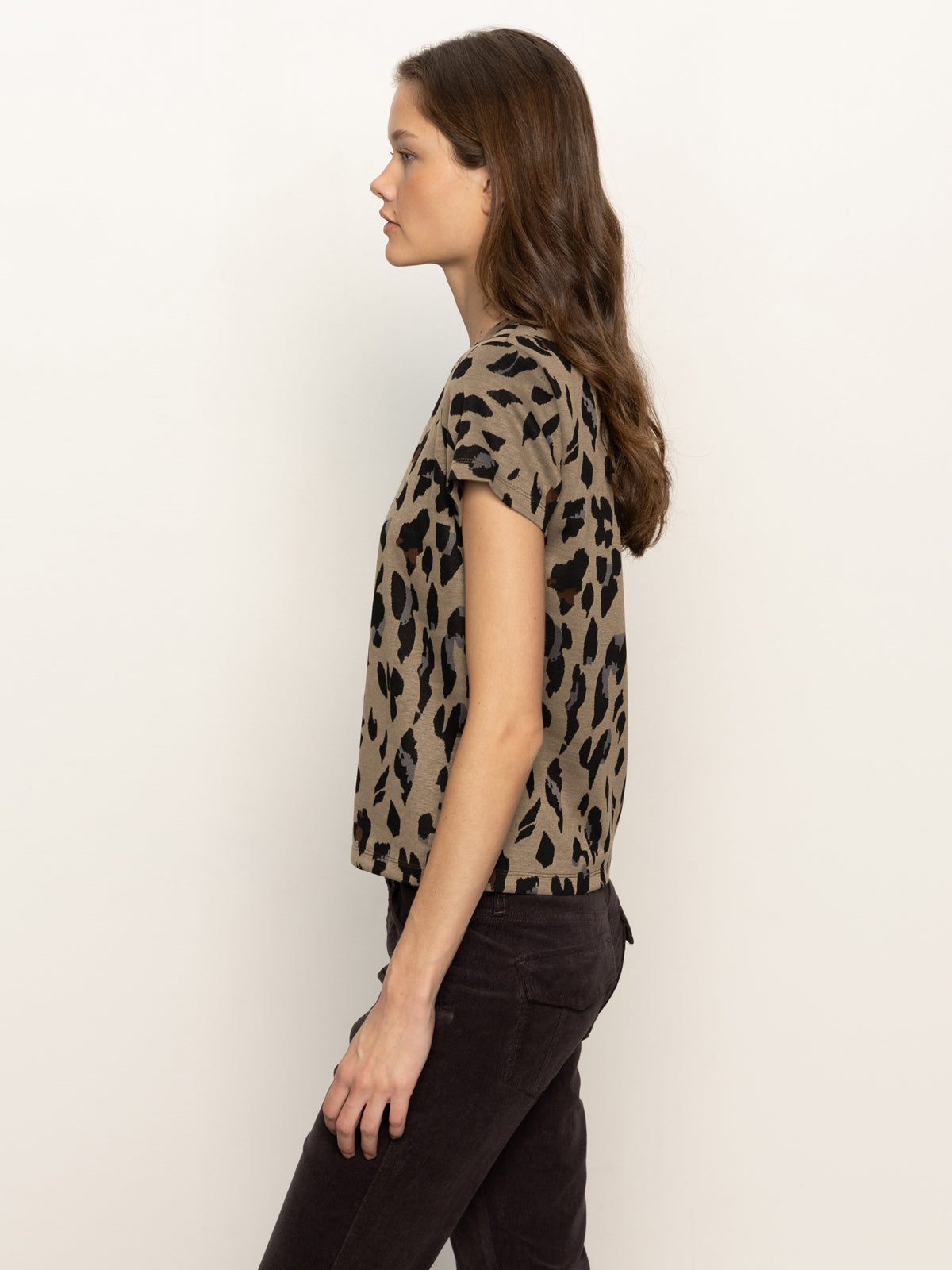 A woman stands in profile against a light background, wearing Sanctuary Clothing’s double knit perfect tee winter leopard—a short-sleeved, beige top with a black leopard print pattern—and dark pants. Her loose brown hair gives her a calm, relaxed look.