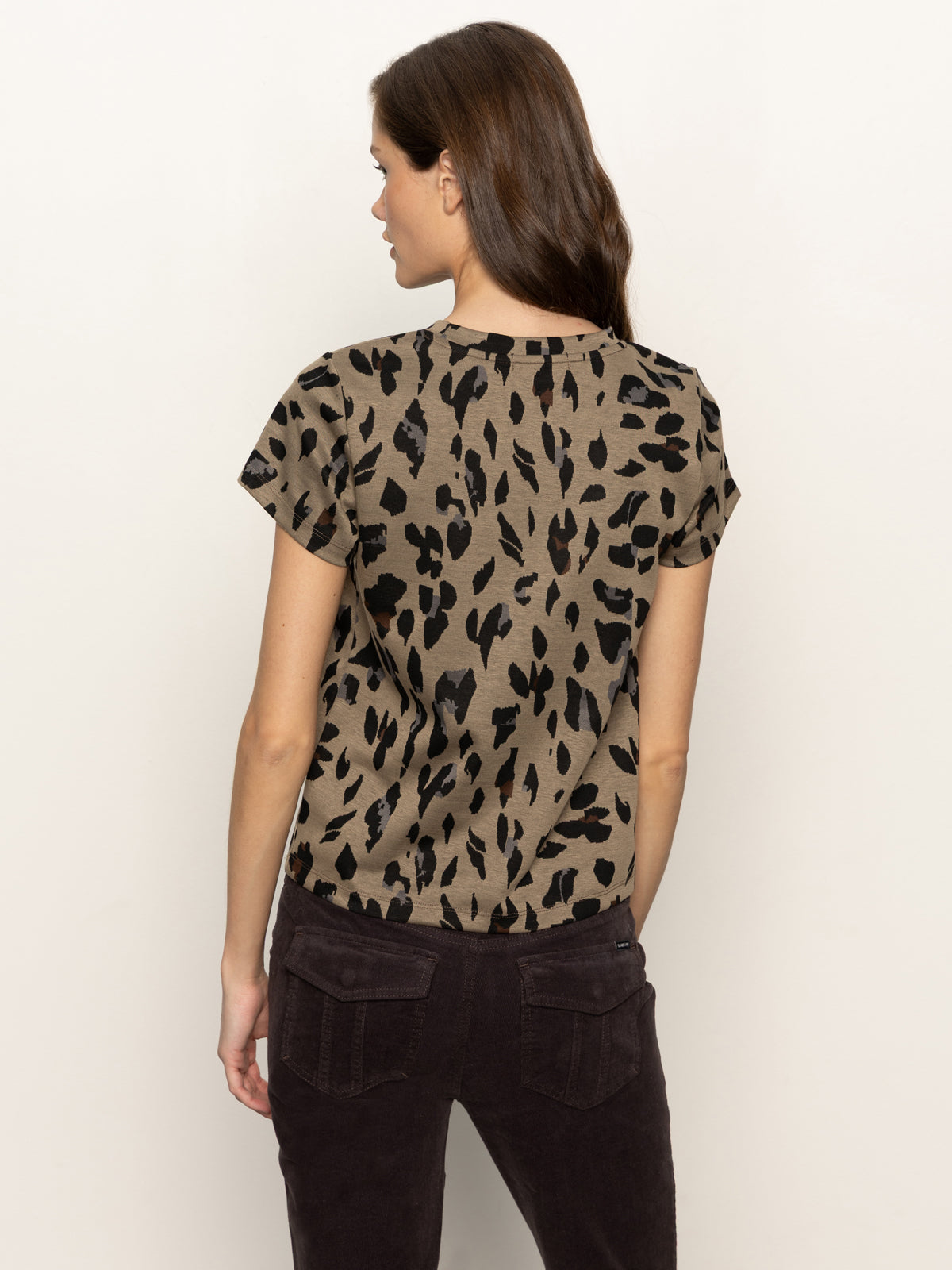 A woman with long brown hair, seen from behind, wears the Sanctuary Clothing double knit perfect tee winter leopard—a short-sleeved brown leopard print top—paired with dark pants against a plain light background.