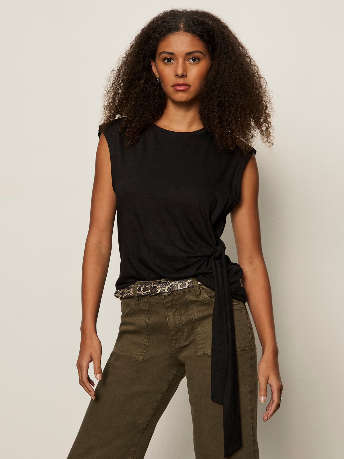A woman with curly hair wears the Sanctuary Clothing side tie muscle tee in black, styled with olive green pants and a patterned belt, standing against a plain light background.